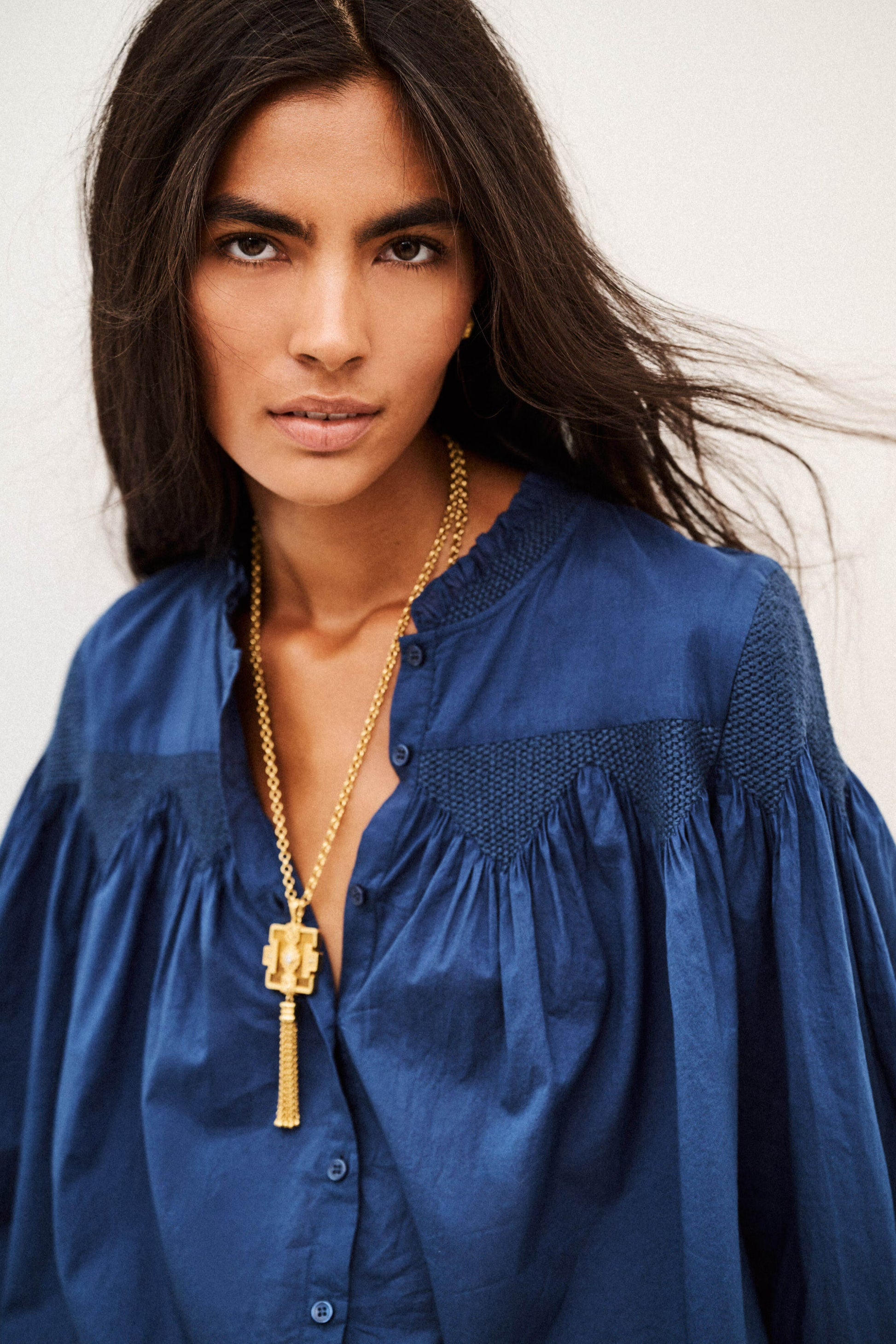 A woman wearing a blue blouse and a gold pendant necklace, photographed against a plain light background.