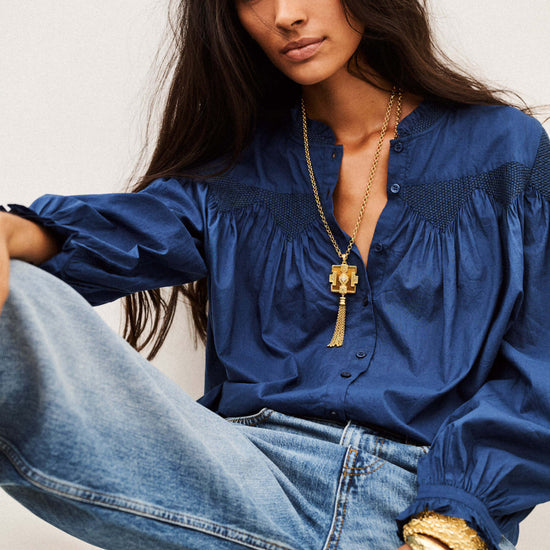 A woman wearing a blue long-sleeve blouse with gathered detailing and a gold pendant necklace, paired with light-wash blue jeans, seated against a plain light background.