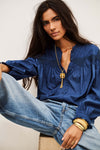 A woman wearing a blue long-sleeve blouse with gathered detailing and a gold pendant necklace, paired with light-wash blue jeans, seated against a plain light background.