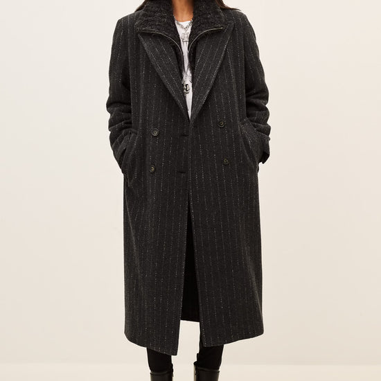 A model wearing a long black double-breasted coat with subtle vertical pinstripes, styled with a white top, black pants, and black boots, set against a plain off-white studio background.