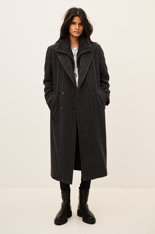 A model wearing a long black double-breasted coat with subtle vertical pinstripes, styled with a white top, black pants, and black boots, set against a plain off-white studio background.
