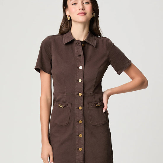 A woman modeling a short-sleeve brown shirt dress with gold-tone buttons and flap pockets, photographed against a light gray studio background.