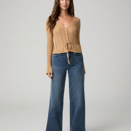 A woman modeling a beige ribbed knit long-sleeve top with a tie-front detail and blue wide-leg jeans against a light gray studio background.