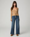 A woman modeling a beige ribbed knit long-sleeve top with a tie-front detail and blue wide-leg jeans against a light gray studio background.