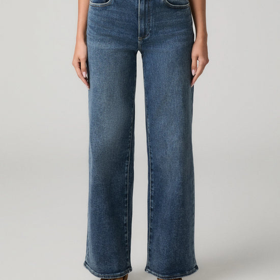A model wearing a pair of high-waisted, wide-leg blue denim jeans with brown suede boots, shown from the waist down against a light gray studio background.