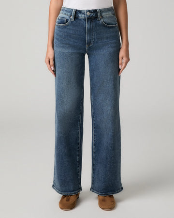A model wearing a pair of high-waisted, wide-leg blue denim jeans with brown suede boots, shown from the waist down against a light gray studio background.