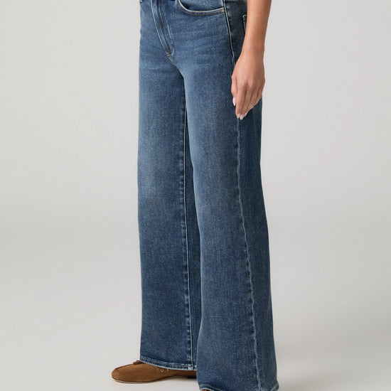 A person wearing blue wide-leg denim jeans with a white top and brown flat shoes, shown from the waist down against a light gray studio background.