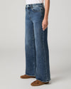 A person wearing blue wide-leg denim jeans with a white top and brown flat shoes, shown from the waist down against a light gray studio background.
