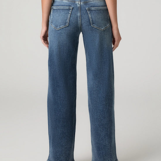 Rear view of a person wearing blue wide-leg jeans with a white top, standing against a light gray studio background.