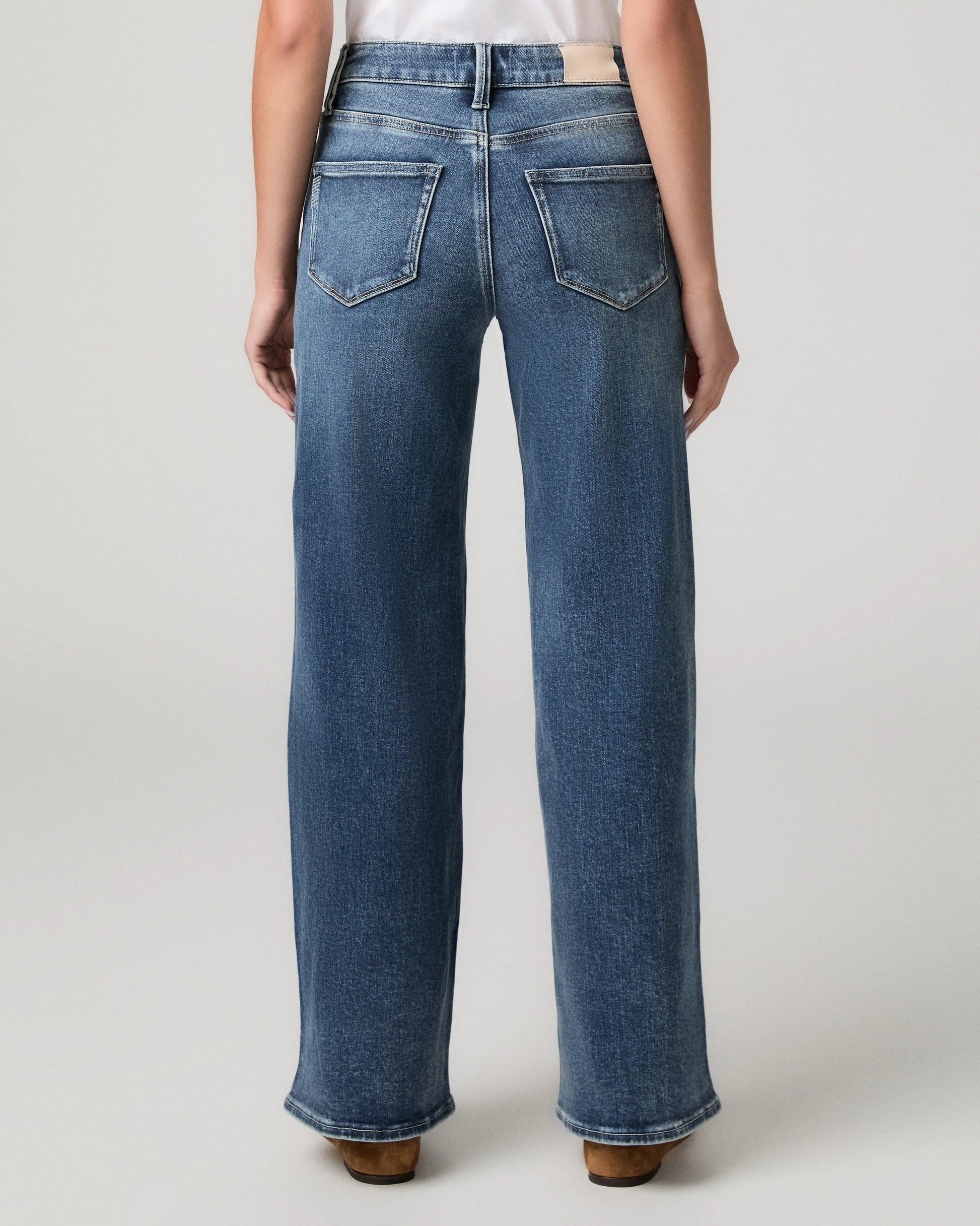 Rear view of a person wearing blue wide-leg jeans with a white top, standing against a light gray studio background.