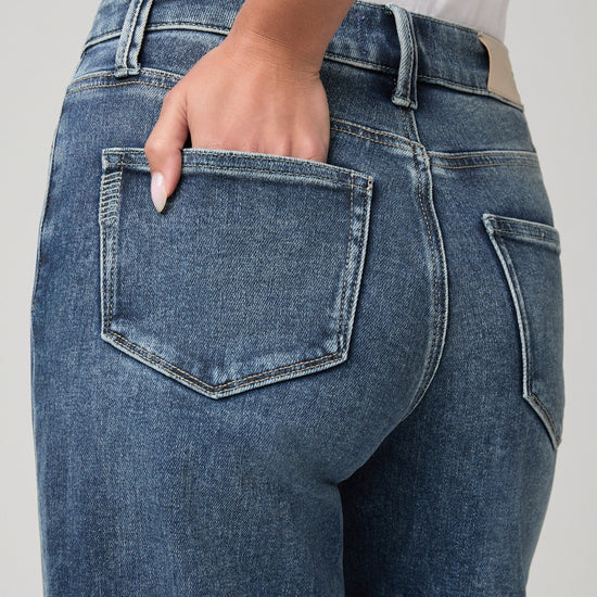 Close-up back view of a person wearing medium-wash blue denim jeans, with one hand tucked into the back pocket.
