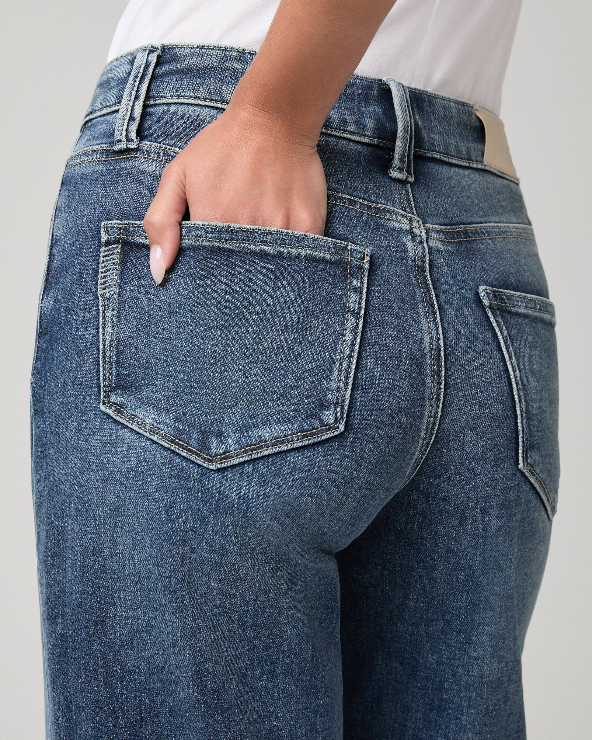 Close-up back view of a person wearing medium-wash blue denim jeans, with one hand tucked into the back pocket.