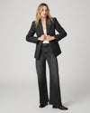 A woman modeling a dark gray textured blazer, dark gray wide-leg jeans, a white top, and black boots against a light gray studio background.