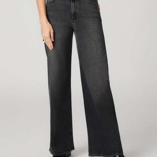 A model wearing a pair of wide-leg dark gray denim jeans with a faded wash, paired with black leather boots against a light gray studio background.