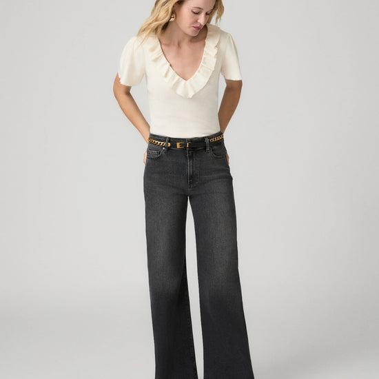A woman modeling a cream short-sleeve top with a ruffled neckline and dark charcoal wide-leg jeans with a gold-tone studded belt, standing against a light gray studio background.