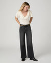 A woman modeling a cream short-sleeve top with a ruffled neckline and dark charcoal wide-leg jeans with a gold-tone studded belt, standing against a light gray studio background.