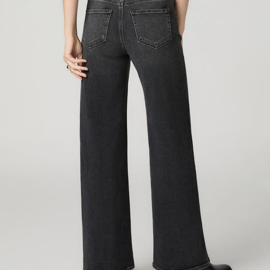 Rear view of a model wearing dark gray wide-leg jeans with a faded wash, paired with a white top and black shoes, against a light gray studio background.