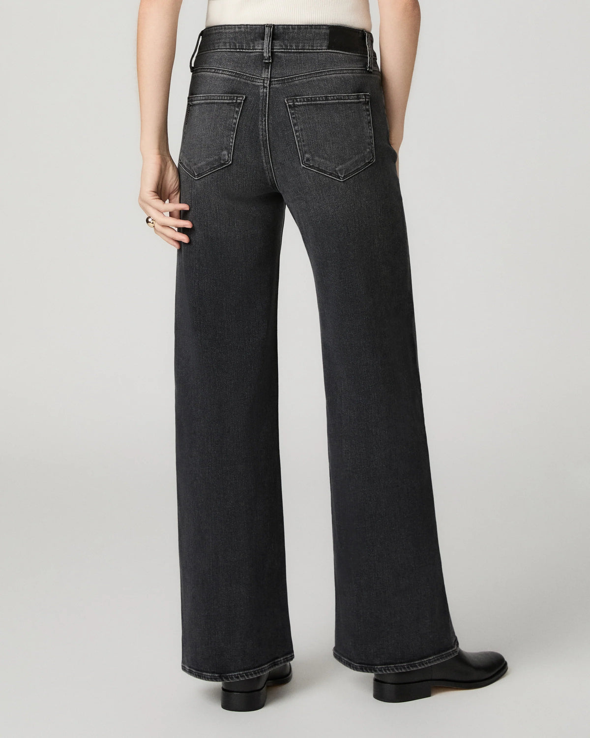 Rear view of a model wearing dark gray wide-leg jeans with a faded wash, paired with a white top and black shoes, against a light gray studio background.