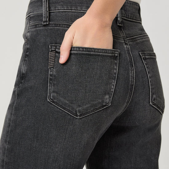 Close-up back view of a person wearing dark charcoal gray denim jeans, with one hand tucked into the back pocket against a light gray studio background.