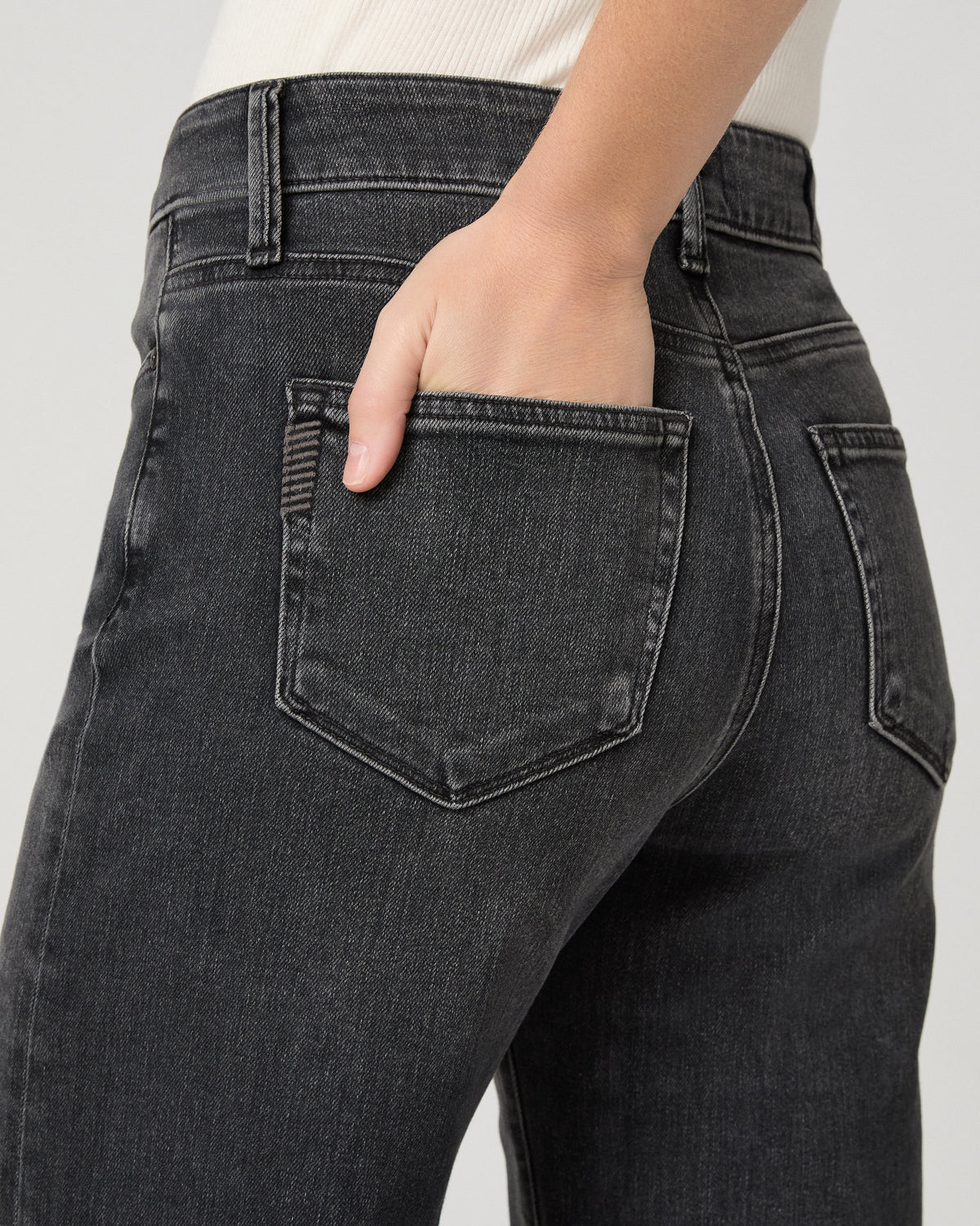 Close-up back view of a person wearing dark charcoal gray denim jeans, with one hand tucked into the back pocket against a light gray studio background.