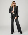 A woman modeling a dark gray textured blazer with wide lapels, paired with dark gray wide-leg jeans, a cream top, and black shoes, against a light gray studio background.