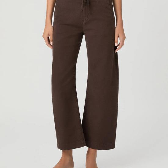 A model wearing dark brown wide-leg cropped pants with a drawstring waist, paired with brown slide sandals, against a light gray studio background.