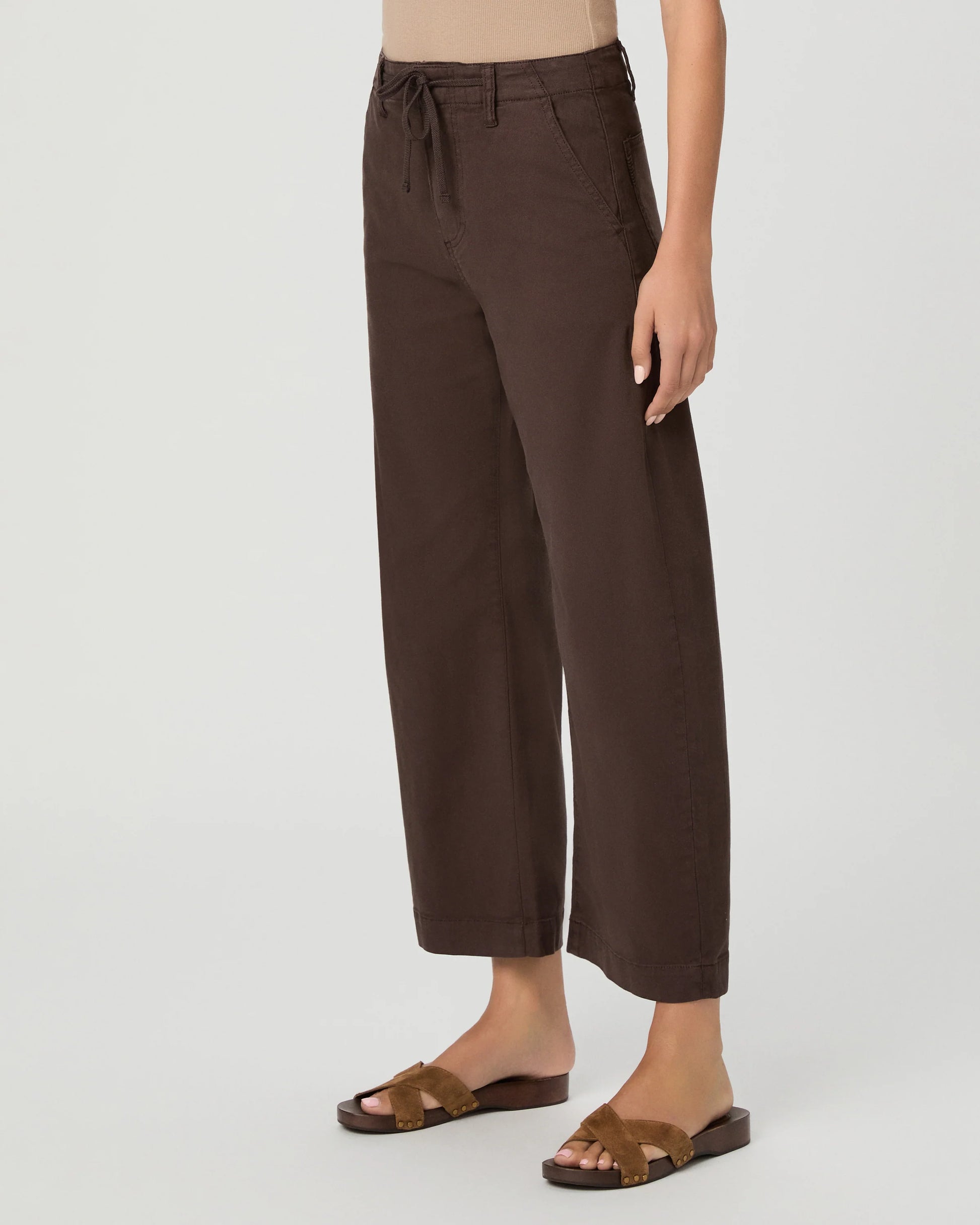 A model wearing dark brown wide-leg cropped pants with a drawstring waistband, paired with brown slide sandals, against a light gray studio background.