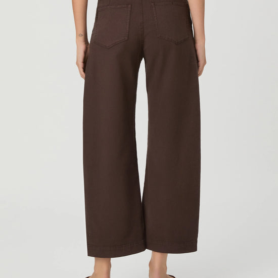 A person shown from the back wearing dark brown wide-leg cropped pants with back patch pockets, paired with tan slide sandals, against a light gray studio background.
