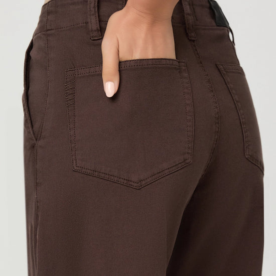 Close-up of a person wearing dark brown wide-leg pants, showing the back pocket and hand in the pocket, against a light gray background.