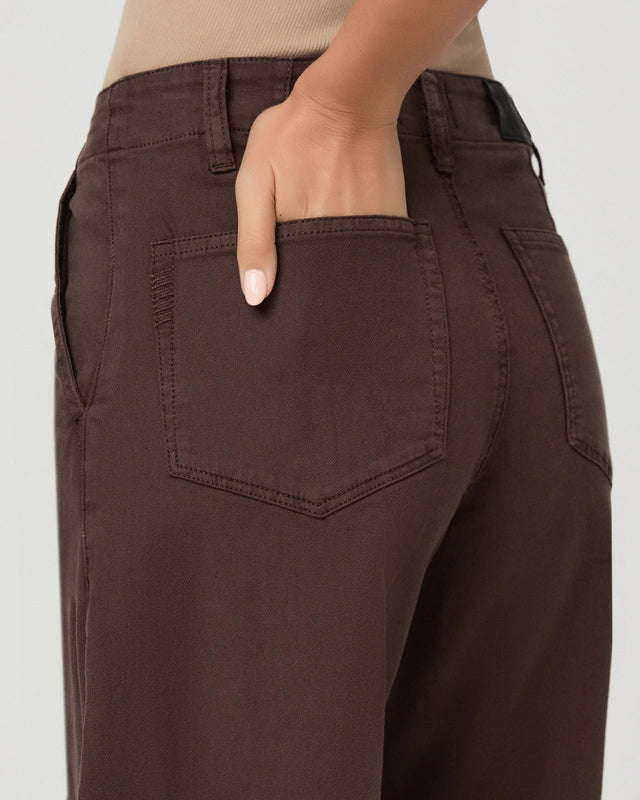 Close-up of a person wearing dark brown wide-leg pants, showing the back pocket and hand in the pocket, against a light gray background.