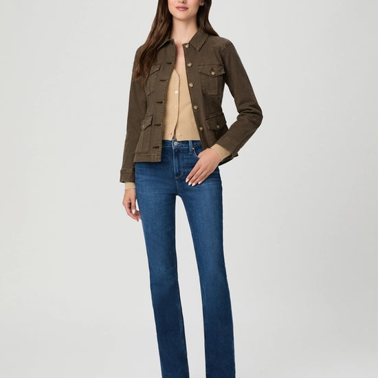 A woman modeling a dark brown utility jacket over a beige top and blue jeans, standing against a light gray studio background.