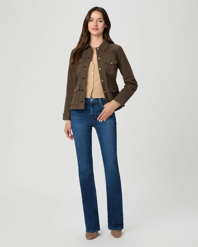 A woman modeling a dark brown utility jacket over a beige top and blue jeans, standing against a light gray studio background.