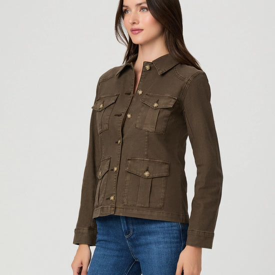 A woman modeling a dark brown utility jacket with four front flap pockets and brass buttons, paired with blue jeans, against a light gray studio background.