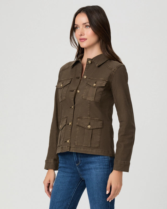 A woman modeling a dark brown utility jacket with four front flap pockets and brass buttons, paired with blue jeans, against a light gray studio background.
