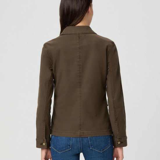 A woman viewed from the back wearing an olive brown utility jacket and blue jeans against a light gray studio background.