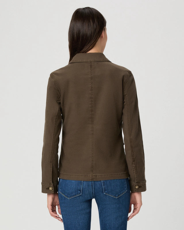 A woman viewed from the back wearing an olive brown utility jacket and blue jeans against a light gray studio background.