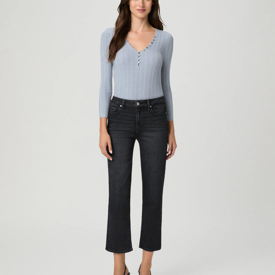 A woman modeling a light blue ribbed knit long-sleeve top with decorative buttons down the front, paired with dark cropped jeans and black heels, against a light gray studio background.