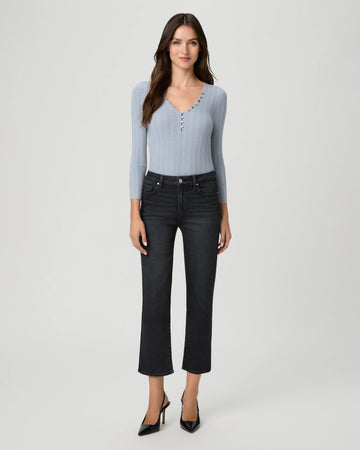A woman modeling a light blue ribbed knit long-sleeve top with decorative buttons down the front, paired with dark cropped jeans and black heels, against a light gray studio background.