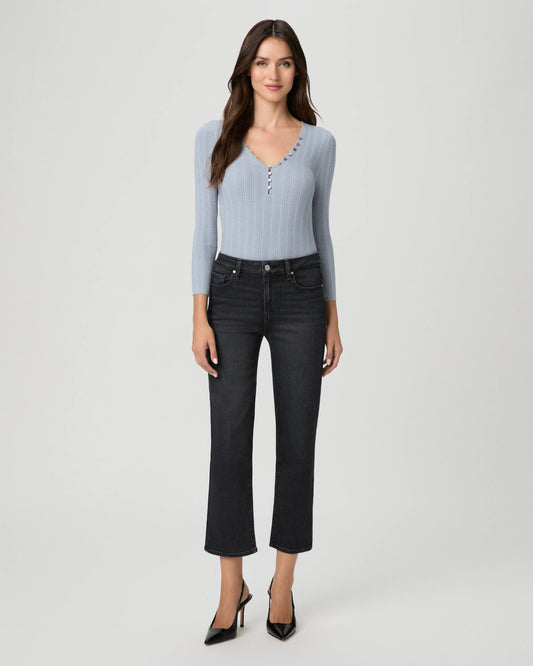 A woman modeling a light blue ribbed knit long-sleeve top with decorative buttons down the front, paired with dark cropped jeans and black heels, against a light gray studio background.