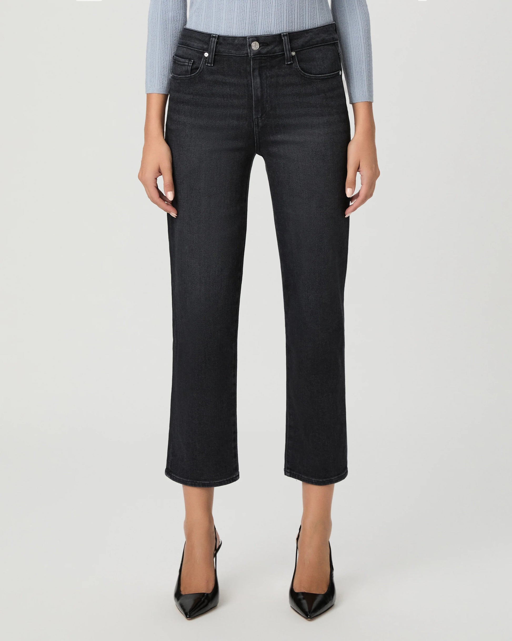 A model wearing a pair of dark charcoal gray cropped jeans, styled with a light blue ribbed long-sleeve top and black pointed-toe heels, against a light gray studio background.