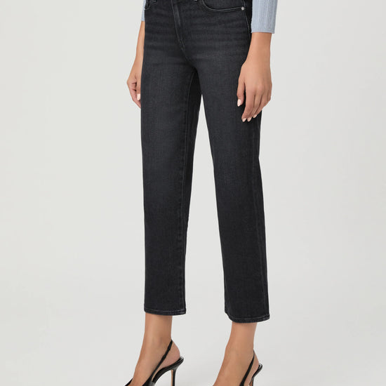 A model wearing a pair of dark charcoal gray cropped jeans with a straight-leg fit, paired with black slingback heels and a light blue striped top, shown against a plain light gray studio background.