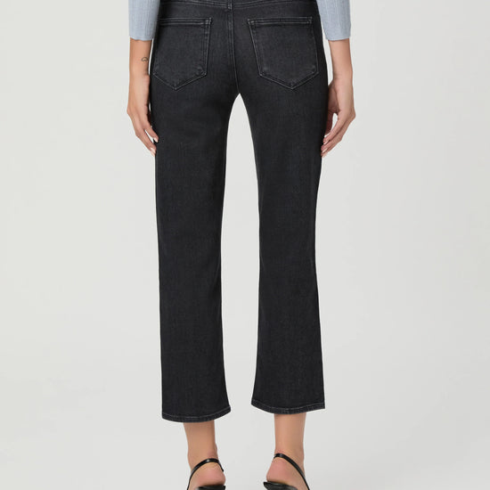 Rear view of a model wearing dark charcoal gray cropped jeans with a straight-leg fit, paired with black high-heeled sandals and a light blue long-sleeve top, against a light gray studio background.