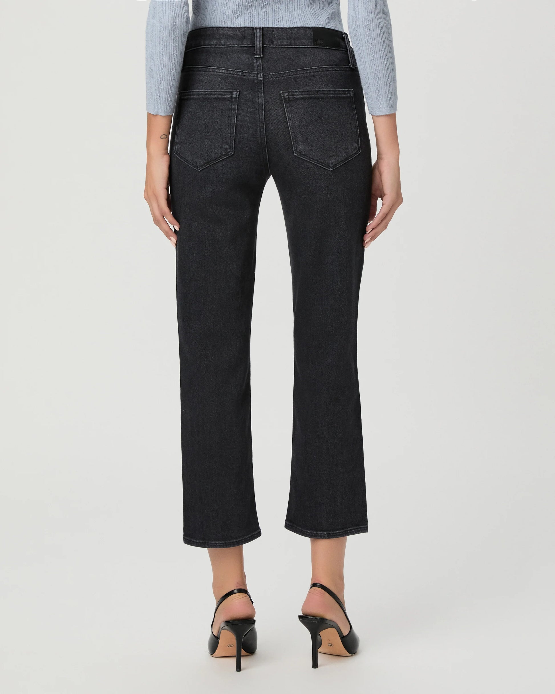 Rear view of a model wearing dark charcoal gray cropped jeans with a straight-leg fit, paired with black high-heeled sandals and a light blue long-sleeve top, against a light gray studio background.
