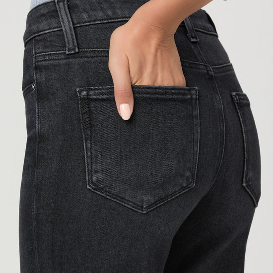 Close-up back view of a person wearing dark charcoal gray jeans, with one hand tucked into the back pocket. A light blue ribbed top is partially visible at the top edge against a plain light gray background.