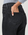 Close-up back view of a person wearing dark charcoal gray jeans, with one hand tucked into the back pocket. A light blue ribbed top is partially visible at the top edge against a plain light gray background.