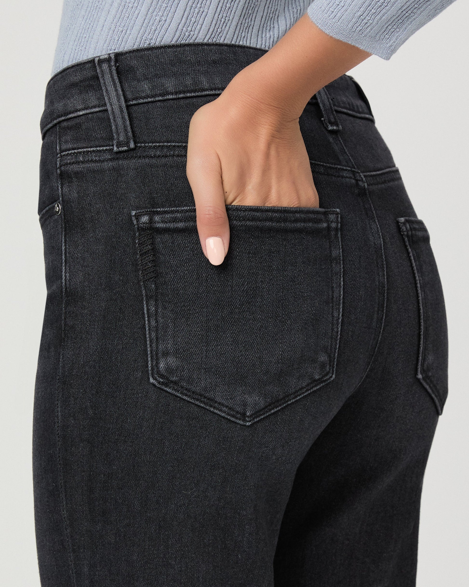 Close-up back view of a person wearing dark charcoal gray jeans, with one hand tucked into the back pocket. A light blue ribbed top is partially visible at the top edge against a plain light gray background.