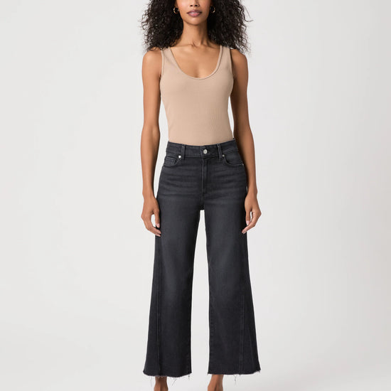 A model stands against a light gray studio background wearing a beige ribbed tank top, dark charcoal wide-leg cropped jeans, and black sandals.