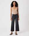 A model stands against a light gray studio background wearing a beige ribbed tank top, dark charcoal wide-leg cropped jeans, and black sandals.