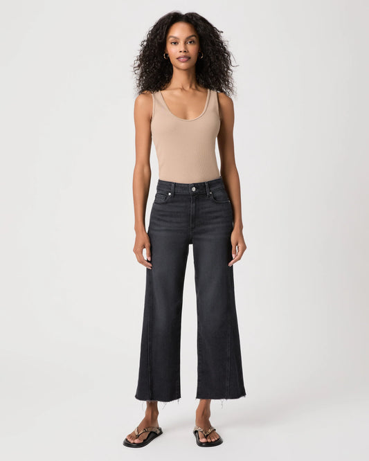 A model stands against a light gray studio background wearing a beige ribbed tank top, dark charcoal wide-leg cropped jeans, and black sandals.