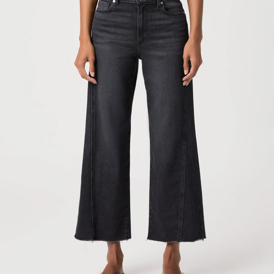 A model wearing a pair of dark charcoal gray wide-leg cropped jeans with a raw hem, paired with black slide sandals, shown against a plain light gray studio background.